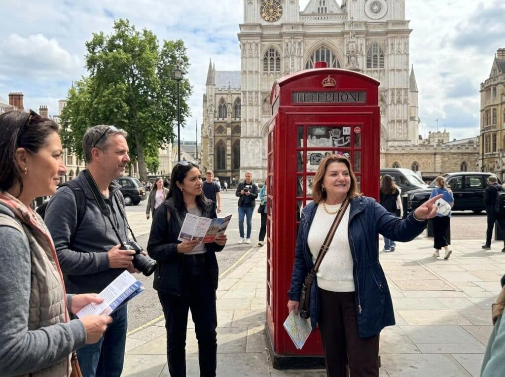 Tour group with maps and brochures stands near a red British phone booth in front of Westminster Abbey in London.